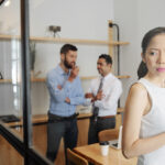 Serious young businesswoman eavesdropping on conversation of her male colleagues in meeting room