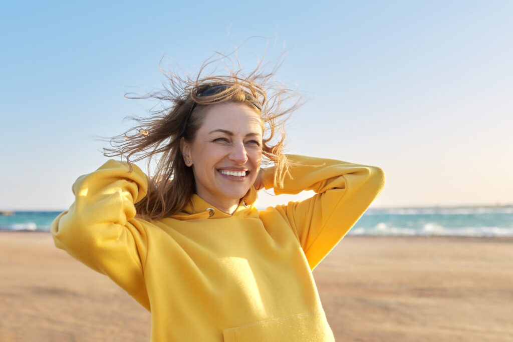 Outdoor portrait of smiling happy blonde woman 45 years old looking at camera. Sunny day on sea coast, beach blue sky background