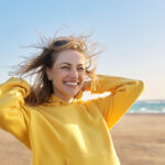 Outdoor portrait of smiling happy blonde woman 45 years old looking at camera. Sunny day on sea coast, beach blue sky background