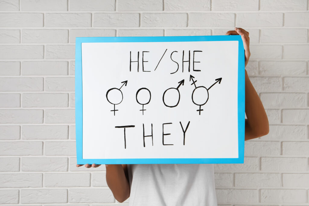 Woman holding sign with gender pronouns and symbols near white brick wall