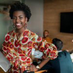 Beautiful young smiling professional black african business woman holding laptop, coworkers hold a meeting in background