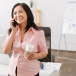 Glad to hear you. Cheerful pregnant asian woman talking on smart phone and drinking coffee while standing in the office
