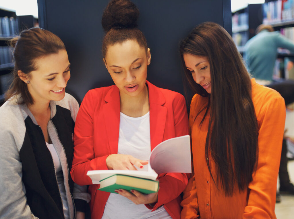 Three young female students standing in the library and looking at a book together. University students reading reference books for their studies.