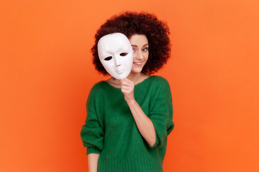 Woman with Afro hairstyle in green sweater removing white mask from face showing his smiling expression, good mood, pretending to be another person. Indoor studio shot isolated on orange background.